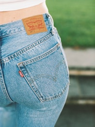 A close-up view of a person wearing a pair of blue Levi Strauss & Co. jeans, showcasing the iconic brown leather label on the waistband and a red tag on the pocket. The background features a blurred green area, possibly grass.