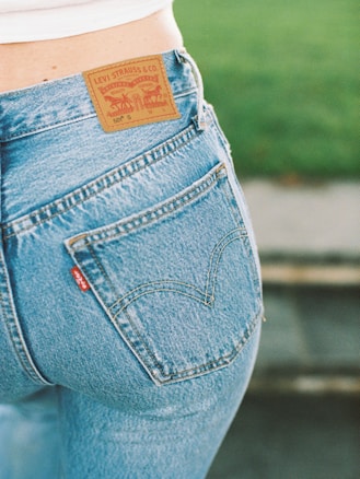 A close-up view of a person wearing a pair of blue Levi Strauss & Co. jeans, showcasing the iconic brown leather label on the waistband and a red tag on the pocket. The background features a blurred green area, possibly grass.