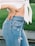 A close-up of a person wearing high-waisted, light blue denim jeans with distressed details and a white, knotted shirt. The person has one hand on their waist, emphasizing casual style and comfort while standing on a grassy background.