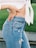 A close-up of a person wearing high-waisted, light blue denim jeans with distressed details and a white, knotted shirt. The person has one hand on their waist, emphasizing casual style and comfort while standing on a grassy background.