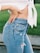 A close-up of a person wearing high-waisted, light blue denim jeans with distressed details and a white, knotted shirt. The person has one hand on their waist, emphasizing casual style and comfort while standing on a grassy background.