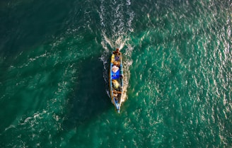 A small fishing boat navigates through vibrant, teal waters, creating ripples as it moves. The boat is filled with fishing gear and a few people, with its bright colors contrasting against the sea.