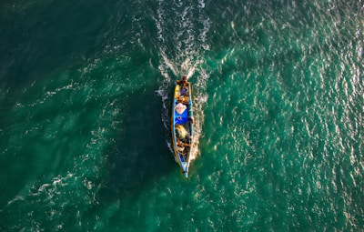 A small fishing boat navigates through vibrant, teal waters, creating ripples as it moves. The boat is filled with fishing gear and a few people, with its bright colors contrasting against the sea.