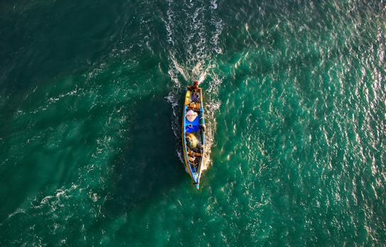 A small fishing boat navigates through vibrant, teal waters, creating ripples as it moves. The boat is filled with fishing gear and a few people, with its bright colors contrasting against the sea.