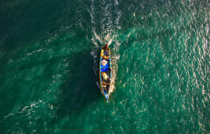 A small fishing boat navigates through vibrant, teal waters, creating ripples as it moves. The boat is filled with fishing gear and a few people, with its bright colors contrasting against the sea.