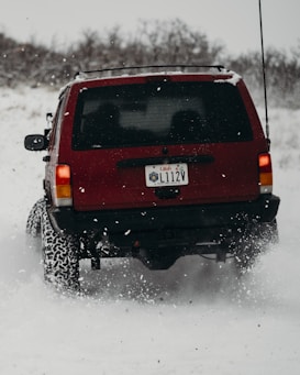 A red SUV is driving on a snowy path, kicking up snow behind it. The vehicle has a Utah license plate and appears to be navigating through a winter landscape. Snow-covered trees are visible in the background.