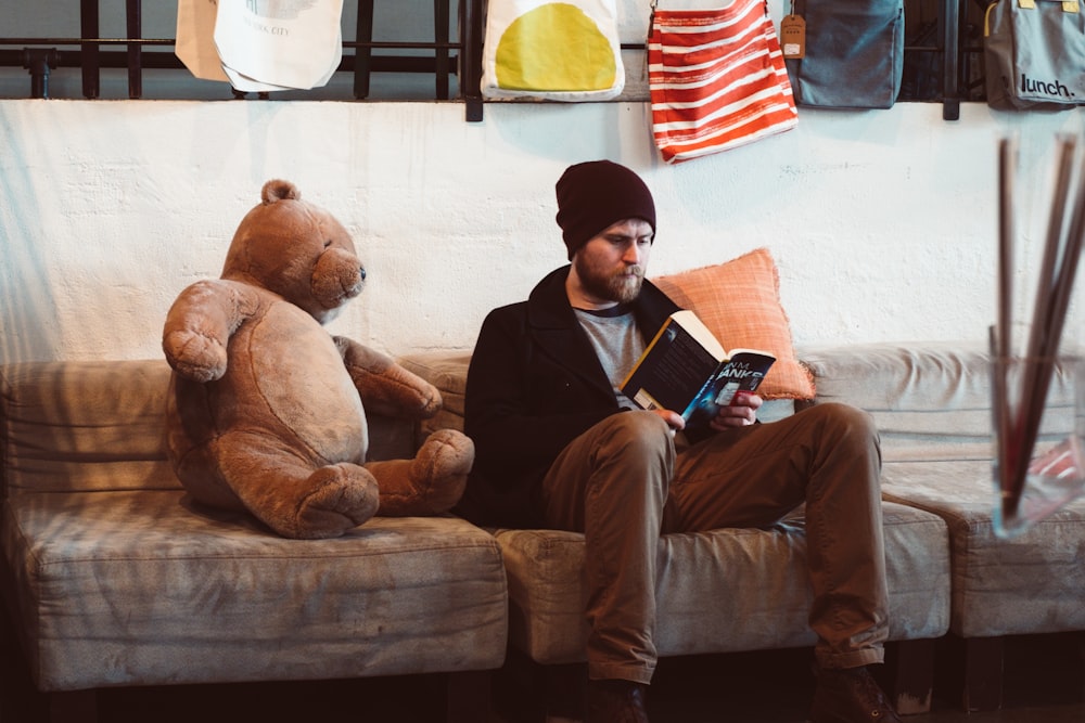 man sitting on sofa reading book near brown bear plush toy