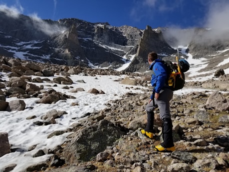 A person wearing a blue jacket and climbing gear is making their way across a rocky, snow-covered mountainous landscape. The backdrop features steep rocky cliffs and a clear blue sky with some clouds. The person appears to be engaged in a hiking or mountaineering activity.
