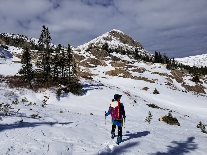 A person wearing a backpack and winter clothing is hiking or trekking through a snowy mountainous landscape. The foreground features a mix of snow-covered ground and scattered rocks, while the background showcases a prominent mountain peak and clusters of evergreen trees. The sky is mostly cloudy, adding depth to the scene.
