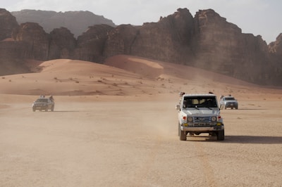 Three off-road vehicles drive across a vast, arid desert landscape with large rocky formations in the background. The sand is light orange and appears dry and untouched aside from the tracks being left by the vehicles. There are a few people visible, standing through the sunroofs of the vehicles.