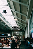 A bustling indoor market with high ceilings and exposed beams. Numerous people are seated at long tables, engaging in conversation and enjoying food and drinks. Several food stalls with bright signs are visible, offering various cuisines. The market has a cozy and lively atmosphere.