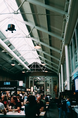 A bustling indoor market with high ceilings and exposed beams. Numerous people are seated at long tables, engaging in conversation and enjoying food and drinks. Several food stalls with bright signs are visible, offering various cuisines. The market has a cozy and lively atmosphere.
