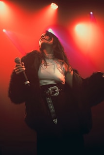 A performer holding a microphone stands under vibrant stage lighting. The lighting creates a dramatic effect, highlighting the figure in a fur-like coat with a decorative belt. The person appears to be immersed in their performance, with a sense of movement and energy.