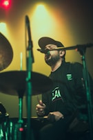 A musician wearing a black cap and jacket is passionately singing into a microphone while playing the drums. The background features colorful, hazy stage lights with an emphasis on green and yellow hues. The scene captures the energy and intensity of a live performance.