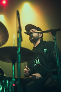 A musician wearing a black cap and jacket is passionately singing into a microphone while playing the drums. The background features colorful, hazy stage lights with an emphasis on green and yellow hues. The scene captures the energy and intensity of a live performance.