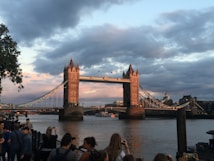 Tower Bridge spans the River Thames under a sky filled with dramatic clouds at dusk. The bridge's towers and suspension sections are illuminated by the warm glow of the setting sun. People are gathered at the riverside, many taking photos of the iconic structure.