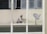 A person wearing protective clothing is painting the exterior of a building while crouched on a narrow ledge. Next to the person is a satellite dish attached to the wall. Several windows are open above the person, providing some ventilation.
