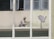 A person wearing protective clothing is painting the exterior of a building while crouched on a narrow ledge. Next to the person is a satellite dish attached to the wall. Several windows are open above the person, providing some ventilation.