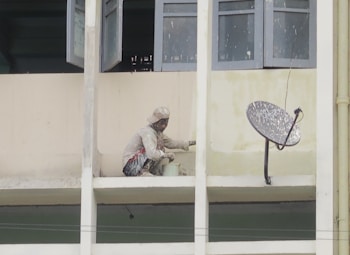A person wearing protective clothing is painting the exterior of a building while crouched on a narrow ledge. Next to the person is a satellite dish attached to the wall. Several windows are open above the person, providing some ventilation.