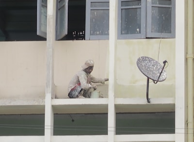 A person wearing protective clothing is painting the exterior of a building while crouched on a narrow ledge. Next to the person is a satellite dish attached to the wall. Several windows are open above the person, providing some ventilation.