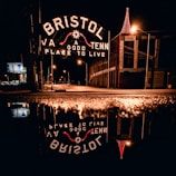 A nighttime scene featuring an illuminated sign reading 'Bristol: A Good Place to Live' with reflections on a wet street surface. The sign spans across the road, and there are streetlights accentuating the urban environment. A building with a brightly lit red Christmas tree or decoration stands on the right.