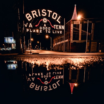 A nighttime scene featuring an illuminated sign reading 'Bristol: A Good Place to Live' with reflections on a wet street surface. The sign spans across the road, and there are streetlights accentuating the urban environment. A building with a brightly lit red Christmas tree or decoration stands on the right.