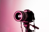 A professional camera mounted on a tripod is highlighted under pink lighting. The focus is on the lens, which has visible brand markings and intricate details that are illuminated against a white background.