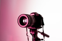 A professional camera mounted on a tripod is highlighted under pink lighting. The focus is on the lens, which has visible brand markings and intricate details that are illuminated against a white background.