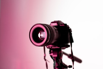 A professional camera mounted on a tripod is highlighted under pink lighting. The focus is on the lens, which has visible brand markings and intricate details that are illuminated against a white background.