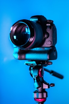 A professional DSLR camera mounted on a sturdy tripod against a solid blue backdrop. The camera is prominently displayed, featuring a large lens and detailed controls, suggesting it's ready for a photoshoot.