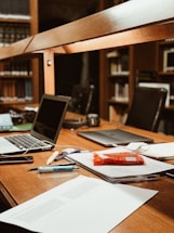 A workspace in a library setting features a wooden desk cluttered with academic materials. Open laptops, scattered papers, highlighters, pens, and a snack pack are visible. Bookshelves filled with books serve as a backdrop, adding to the scholarly atmosphere.
