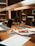 A workspace in a library setting features a wooden desk cluttered with academic materials. Open laptops, scattered papers, highlighters, pens, and a snack pack are visible. Bookshelves filled with books serve as a backdrop, adding to the scholarly atmosphere.