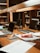 A workspace in a library setting features a wooden desk cluttered with academic materials. Open laptops, scattered papers, highlighters, pens, and a snack pack are visible. Bookshelves filled with books serve as a backdrop, adding to the scholarly atmosphere.