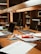 A workspace in a library setting features a wooden desk cluttered with academic materials. Open laptops, scattered papers, highlighters, pens, and a snack pack are visible. Bookshelves filled with books serve as a backdrop, adding to the scholarly atmosphere.