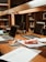 A workspace in a library setting features a wooden desk cluttered with academic materials. Open laptops, scattered papers, highlighters, pens, and a snack pack are visible. Bookshelves filled with books serve as a backdrop, adding to the scholarly atmosphere.