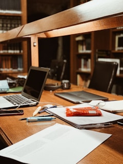 A workspace in a library setting features a wooden desk cluttered with academic materials. Open laptops, scattered papers, highlighters, pens, and a snack pack are visible. Bookshelves filled with books serve as a backdrop, adding to the scholarly atmosphere.