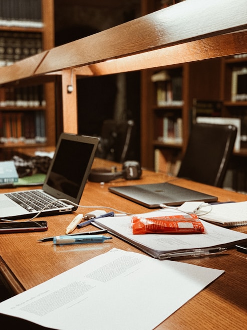 A workspace in a library setting features a wooden desk cluttered with academic materials. Open laptops, scattered papers, highlighters, pens, and a snack pack are visible. Bookshelves filled with books serve as a backdrop, adding to the scholarly atmosphere.