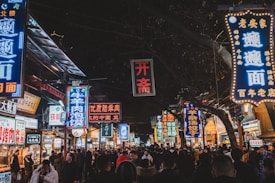 A bustling night market scene with vibrant, glowing signs written in Chinese characters hanging above a crowded street. People walk among the various food stalls and shops, creating a lively and energetic atmosphere. The neon lights illuminate the street, adding a colorful and dynamic touch to the environment.