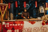 A person wearing a headscarf is preparing snacks at a street food stall. The stall is decorated with traditional Chinese banners and signs, and various snacks are displayed on the counter. The setting appears to be busy, with another customer visible in the background.