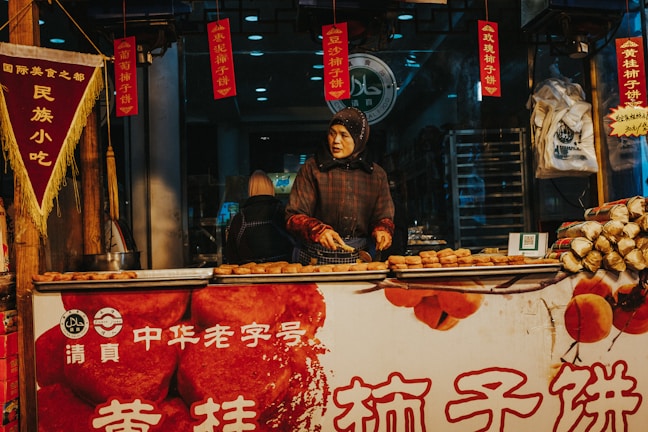 A person wearing a headscarf is preparing snacks at a street food stall. The stall is decorated with traditional Chinese banners and signs, and various snacks are displayed on the counter. The setting appears to be busy, with another customer visible in the background.