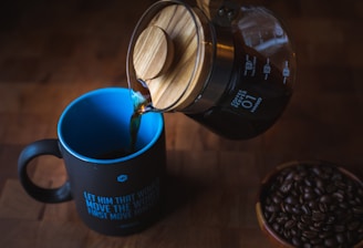 A wooden-topped coffee carafe pours dark coffee into a black mug with a blue interior. The mug has an inspirational quote written on it. Nearby, a bowl is filled with coffee beans. The setting is on a dark wooden surface, creating a warm and cozy atmosphere.