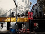A streetscape featuring a pet store with a yellow sign. Several air conditioning units are mounted on the wall above, and two red bicycles are parked under a shaded area. The scene is accented with red signs containing Chinese characters, and there are shadows cast by trees.