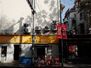A streetscape featuring a pet store with a yellow sign. Several air conditioning units are mounted on the wall above, and two red bicycles are parked under a shaded area. The scene is accented with red signs containing Chinese characters, and there are shadows cast by trees.