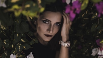 A woman with dark lipstick and piercing eyes peeks through vibrant greenery and flowers, creating a mysterious and captivating atmosphere. Her hand touches her forehead, and she wears a watch on her wrist.