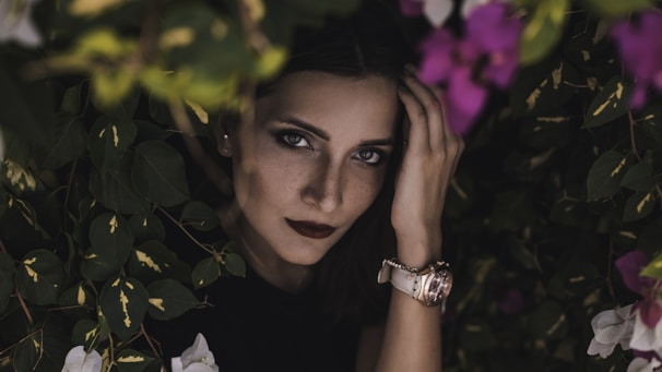 A woman with dark lipstick and piercing eyes peeks through vibrant greenery and flowers, creating a mysterious and captivating atmosphere. Her hand touches her forehead, and she wears a watch on her wrist.