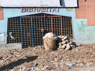 A closed, graffiti-covered storefront with a blue wall and a metal grid gate. Debris is scattered across the dirt ground. Next to the entrance is a large woven basket filled with items and a pile of rocks or cement blocks.