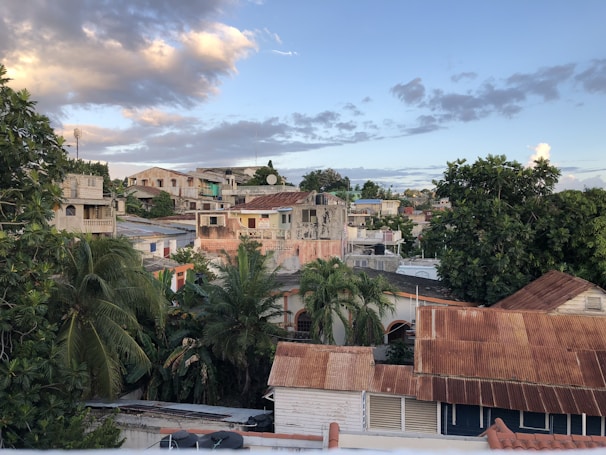 The image depicts a residential area with a mix of old and new buildings. The houses have varied architectural styles, some with rusted metal roofs and others with newer construction. Lush green trees and palms are scattered throughout the neighborhood. The sky above is partly cloudy with the sun lighting up portions of the clouds and landscape.