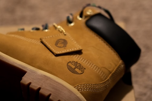 A close-up of a tan leather boot featuring a rugged sole and distinctive stitching. The shoe has a small tag attached by a chain with a logo, and the upper area is padded in black. The focus on detail highlights the texture and branding of the boot.