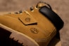 A close-up of a tan leather boot featuring a rugged sole and distinctive stitching. The shoe has a small tag attached by a chain with a logo, and the upper area is padded in black. The focus on detail highlights the texture and branding of the boot.