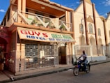 A street-side scene with a two-story building featuring a sign for Guy's Hotel, Guest House, Bar, and Restaurant. The building has a balcony with decorative triangular elements and is painted in earth tones. In front, a person rides a motorcycle on the paved road. Adjacent buildings are visible, one of which has an arched architectural design.
